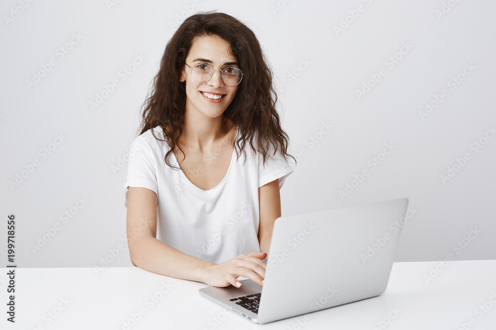 Attractive and smart assistant helping customer to fill out blank. Polite good-looking curly-haired girl in trendy round glasses sitting at table with laptop, typing, working on project over gray wall Attractive and smart assistant helping customer to fill out blank. Polite good-looking curly-haired girl in trendy round glasses sitting at table with laptop, typing, working on project over gray wall