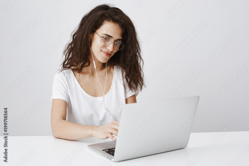 Hard-working girl working under new project, creating concept for company. Focused happy caucasian curly-haired woman typing on laptop, smiling, listening music in earphones, doing home assignment Hard-working girl working under new project, creating concept for company. Focused happy caucasian curly-haired woman typing on laptop, smiling, listening music in earphones, doing home assignment