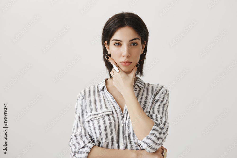 Serious and attractive european girl trying to concentrate while making up new concept for her design, standing over gray background with strict and focused expression, holding hand on jaw, planning Serious and attractive european girl trying to concentrate while making up new concept for her design, standing over gray background with strict and focused expression, holding hand on jaw, planning
