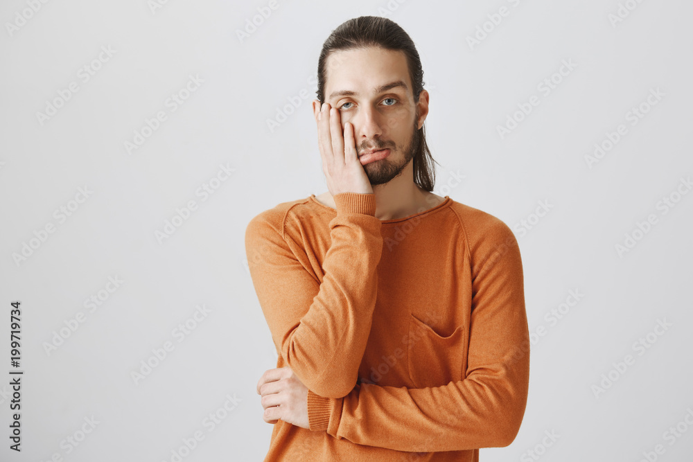 No emotions only facepalm. Portrait of tired and bored attractive guy in casual orange pullover, holding palm on cheek and pouting, being indifferent and uninterested in discussion over gray wall No emotions only facepalm. Portrait of tired and bored attractive guy in casual orange pullover, holding palm on cheek and pouting, being indifferent and uninterested in discussion over gray wall