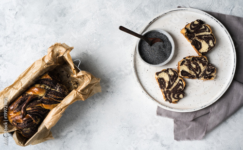 Mohnkuchen geschnitten und hübsch angerichtet  auf weißem Teller und grauem Stofftuch Leinen auf hellem Untergrund mit ganzem Kuchen im Bild von oben fotografiert bei Tageslicht