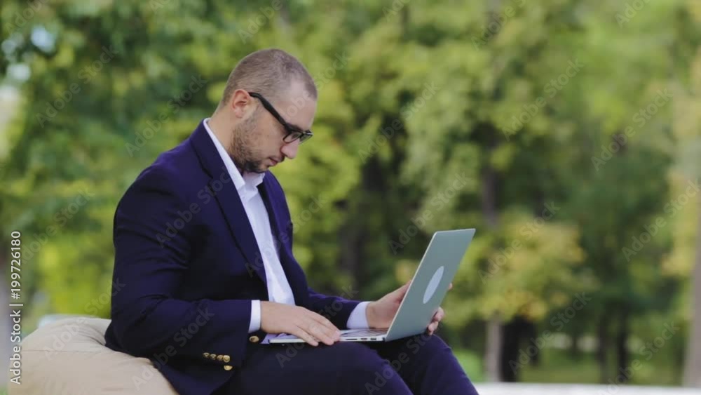 Young disheartened smart man businessman or student in casual blue shirt, glasses sitting at table, prints on a laptop in city park. Crypto investor working outdoors. Crypto-currency operation. Young disheartened smart man businessman or student in casual blue shirt, glasses sitting at table, prints on a laptop in city park. Crypto investor working outdoors. Crypto-currency operation.
