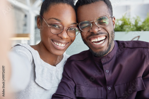Positive dark skinned smiling woman and man have fun together, make selfie, wear glasses, laugh, happy to meet, being good friends, have good friendly relationships. Pleased African American couple