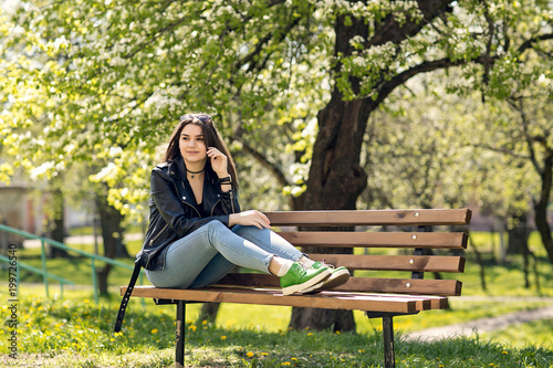 Glamorous young Caucasian woman in black leather jacket sitting in the park on the bench