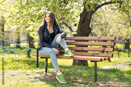 Glamorous young Caucasian woman in black leather jacket sitting in the park on the bench