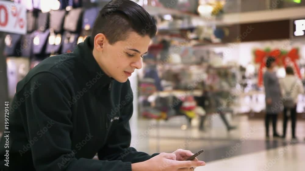Profile of successful young man with crew haircut who sits and keeps a smartphone in his hands. He surfs the net in a shopping mall in autumn Profile of successful young man with crew haircut who sits and keeps a smartphone in his hands. He surfs the net in a shopping mall in autumn