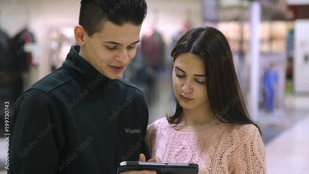 A cheerful view of a young couple standing, smiling and looking at their tablet in a modern shopping mall. They feel happy and curious A cheerful view of a young couple standing, smiling and looking at their tablet in a modern shopping mall. They feel happy and curious
