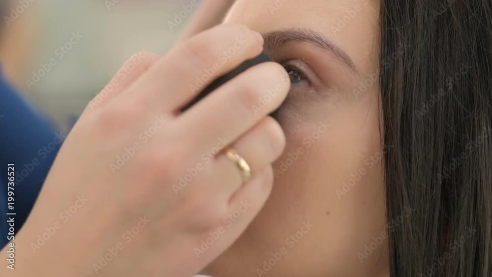 Portrait of young beautiful woman dressed pink sitting in beauty salon and stylist applies tone on cheek with sponge Portrait of young beautiful woman dressed pink sitting in beauty salon and stylist applies tone on cheek with sponge