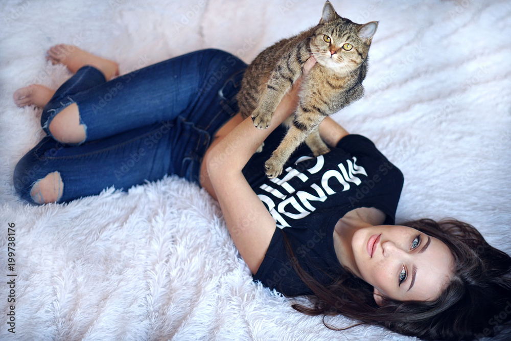 Young brunette woman with long hair and deep blue eyes in torn jeans playing with cat at the bedroom Young brunette woman with long hair and deep blue eyes in torn jeans playing with cat at the bedroom