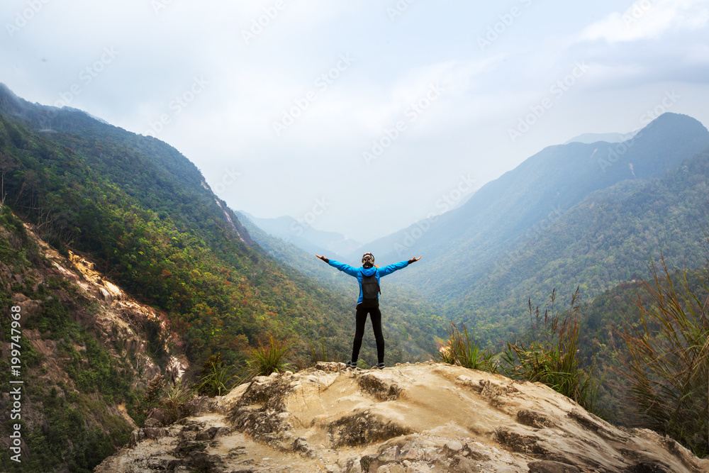 Tourist traveler standing on a rock with raised hands, hiker looking to a valley below in trip in Vietnam, woman enjoying peak of  mountain background landscape