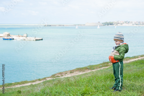 cute pensive little boy in scarf jacket and hat on green grassy slope against sea with floating boats in springtime