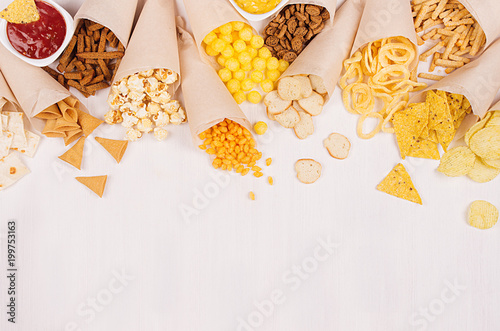 Obraz na plátně Summer fun fast food - different crunchy snacks in craft paper cone and salsa and curry sauces in bowls as decoration border on soft white wood board
