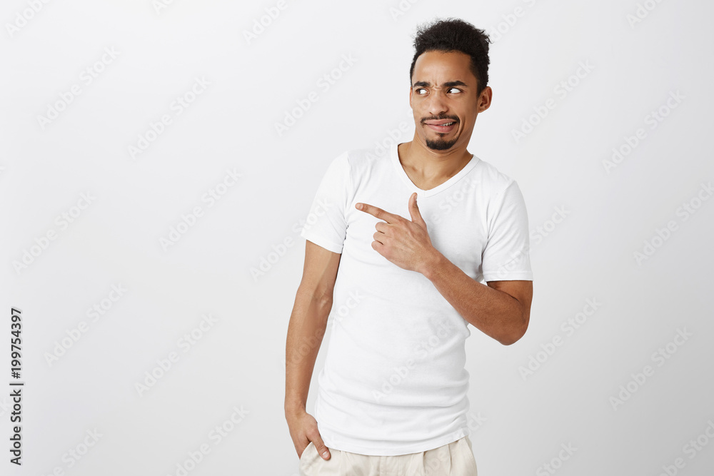 What an ugly thing. Portrait of displeased handsome dark-skinned guy in white t-shirt, tilting from fromething disgusting, pointing left with index finger, expressing disappointment and antipathy What an ugly thing. Portrait of displeased handsome dark-skinned guy in white t-shirt, tilting from fromething disgusting, pointing left with index finger, expressing disappointment and antipathy
