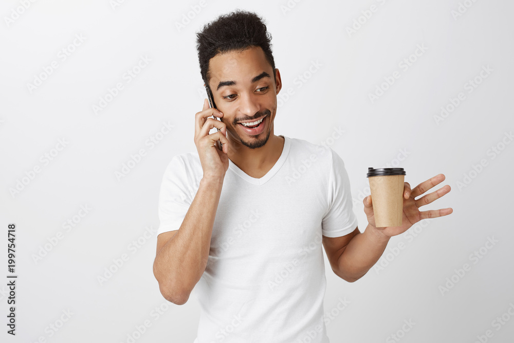 Casual chat during break in cafe. Portrait of happy attractive male model with afro haircut, talking on smartphone with good friend, drinking coffee and gesturing, describing story over gray wall Casual chat during break in cafe. Portrait of happy attractive male model with afro haircut, talking on smartphone with good friend, drinking coffee and gesturing, describing story over gray wall