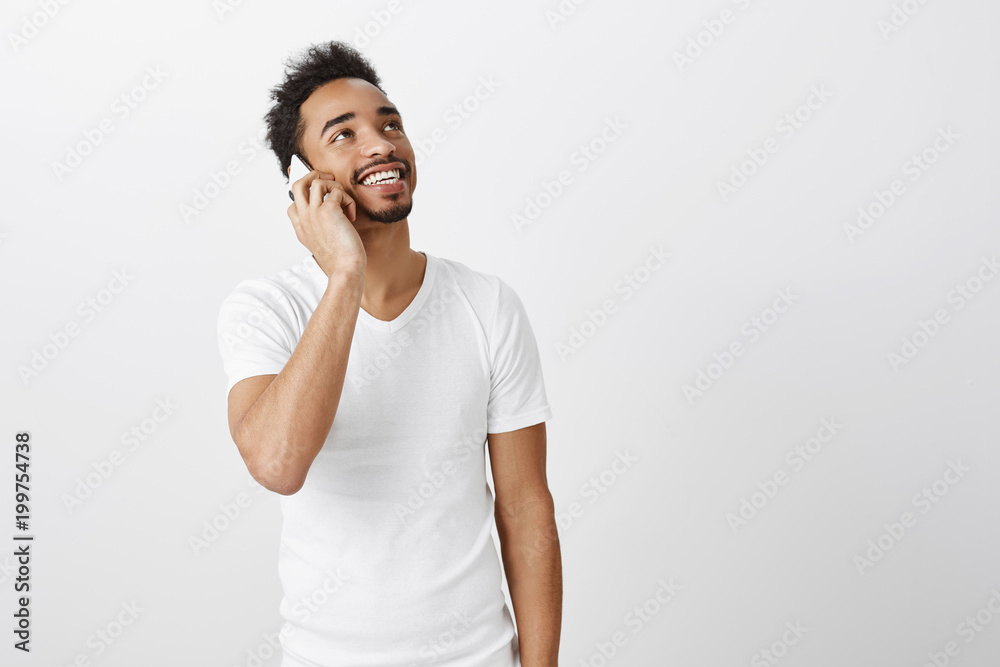 Caring boyfriend enjoys hearing girlfriends voice. Portrait of romantic good-looking dark-skinned male with curly hair, looking up, smiling broadly while talking on smartphone, discussing lifestyle Caring boyfriend enjoys hearing girlfriends voice. Portrait of romantic good-looking dark-skinned male with curly hair, looking up, smiling broadly while talking on smartphone, discussing lifestyle