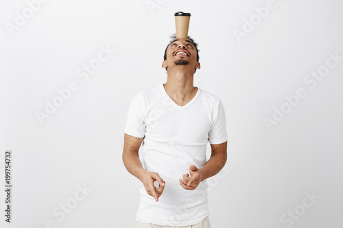 Skillful guy showing his balance. Portrait of good-looking african-american man with beard, tilting head up while holding cup with coffee on forehead, smiling, balancing and fooling around