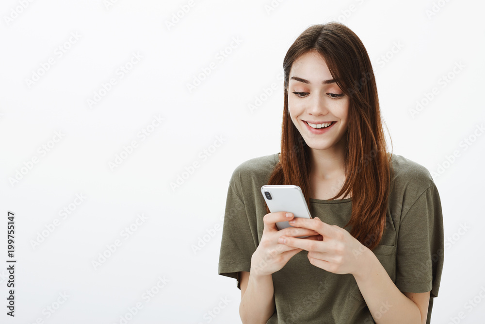 Woman cannot believe in own luck. Studio shot of satisfied good-looking european brunette, holding smartphone and smiling broadly, being excited and fascinated with great message over gray wall Woman cannot believe in own luck. Studio shot of satisfied good-looking european brunette, holding smartphone and smiling broadly, being excited and fascinated with great message over gray wall