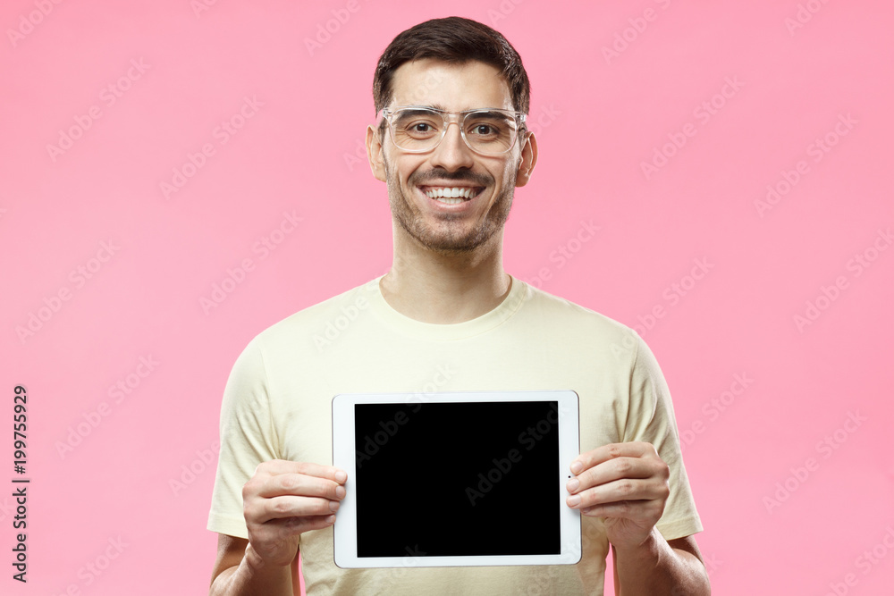 Studio picture of positive man isolated on pink background standing in casual clothes holding tablet and showing it blank screen with happy smile as if advising product or service