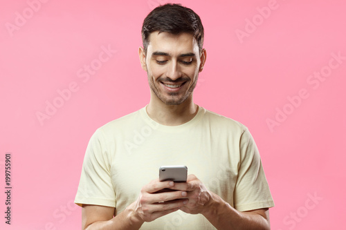 Closeup photo of man in beige tshirt standing isolated on pink background looking attentively at screen of cellphone, browsing web pages and smiling nicely while chatting with friends