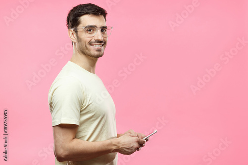 Young attractive man in beige t-shirt looking and smiling at the camera with cheerful expression, using mobile phone, isolated on pink background. Copy space for your advertising text