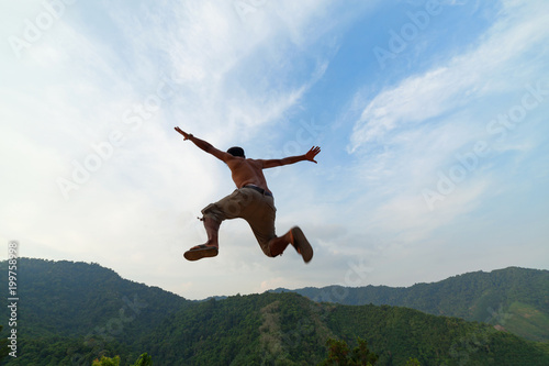 Asian man traveler is jumping on top of a rainforest mountain in scenery background.