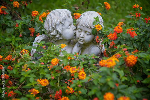 ANGELS IN LOVE. Sculptures of boy and girl angels surrounded with flowers hugging and cuddling. The boy angel is kissing her by the cheek.