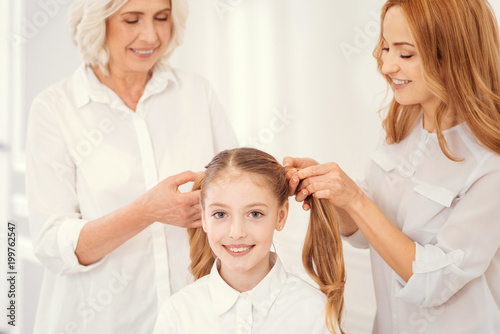 Girly moments. Adorable little girl smiling into the camera while sitting on a chair and waiting for her tender granny and mom making ponytails.