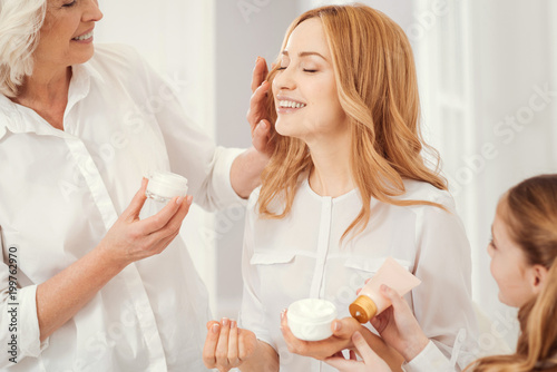 Joyful moment. Radiant mature blonde lady sitting on a chair and beaming while enjoying the time spent with family and taking care of her face skin together with a loveful grandmother and daughter