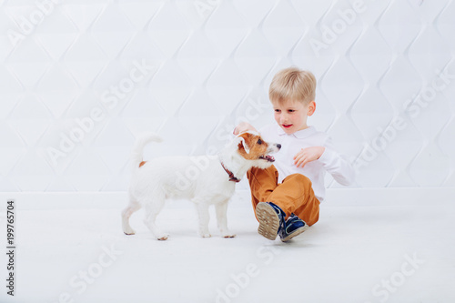 Cute little blond preschooler boy sitting on the floor and playingtogether with a young jack russell terrier dog on a white modern background. Friendship and pet lover concept. photo