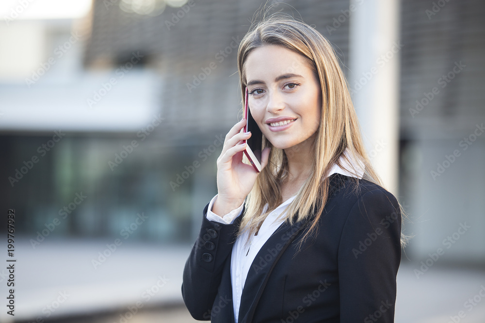 Portrait of smiling business woman talking on phone and looking at the camera. Urban background Portrait of smiling business woman talking on phone and looking at the camera. Urban background