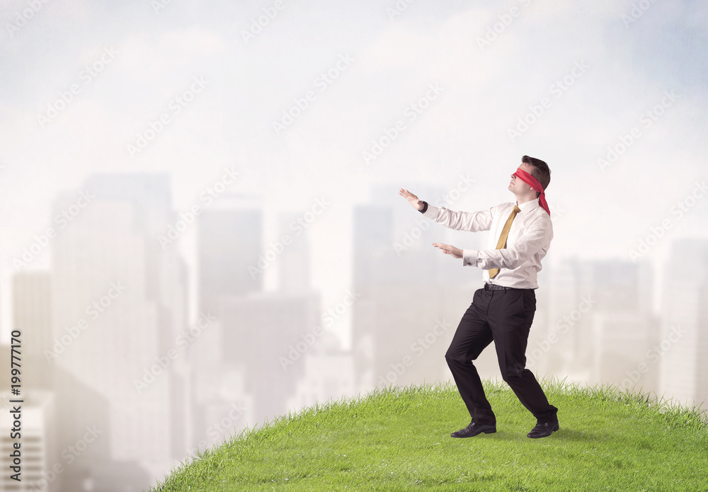 Young blindfolded businessman steps on a a patch of grass with a city in the background Young blindfolded businessman steps on a a patch of grass with a city in the background