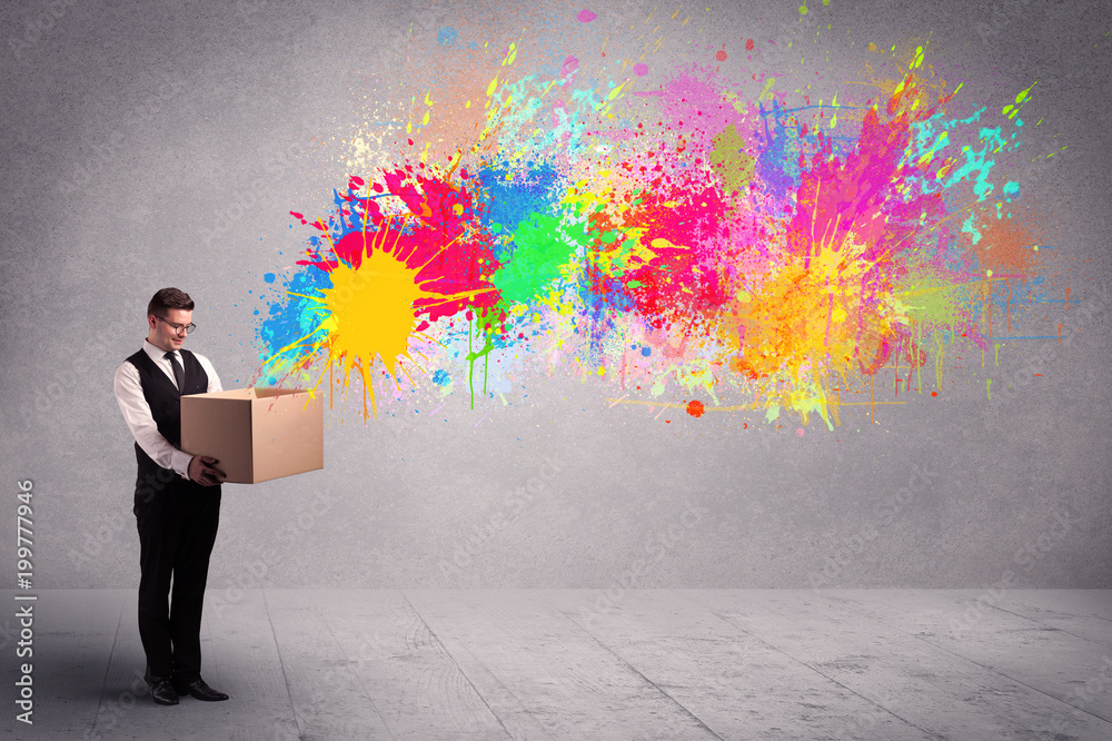 A young smiling business male holding a paperboard box with illustration of colourful spray paint splash on urban wall background concept. A young smiling business male holding a paperboard box with illustration of colourful spray paint splash on urban wall background concept.