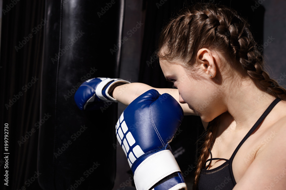 Sideways shot of concentrated 14 year old Caucasian girl with braids wearing black sports bra and blue boxing gloves standing at punch bag, mastering punching technique during training in gym