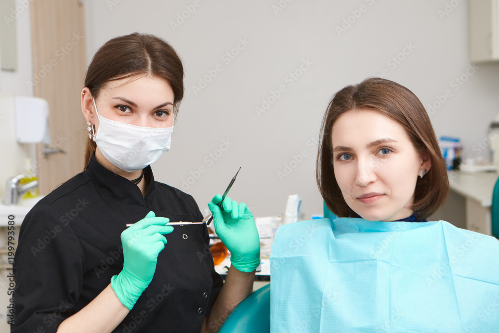 Portrait of attractive confident young female dentist wearing gloves and  mask, holding metal dental probe with small angled mirror, going to examine  oral cavity of woman patient in dentist's chair Stock-Foto