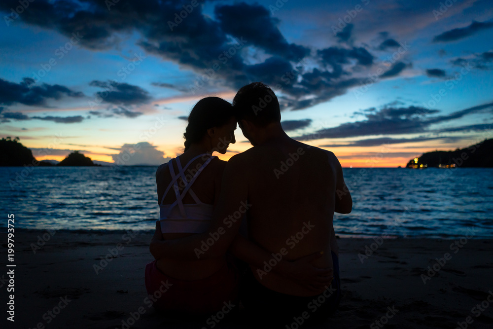 Rear view of a young couple kissing while sitting on a dreamy beach at dusk during summer vacation or honeymoon in Flores Island, Indonesia