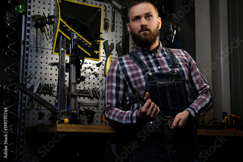Portrait of a handsome stylish male with beard and haircut wearing a flannel shirt and jeans coverall, holds steel wrench, standing in a workshop.