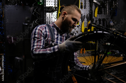 Handsome stylish male wearing a flannel shirt and jeans coverall, working with a bicycle wheel in a repair shop.