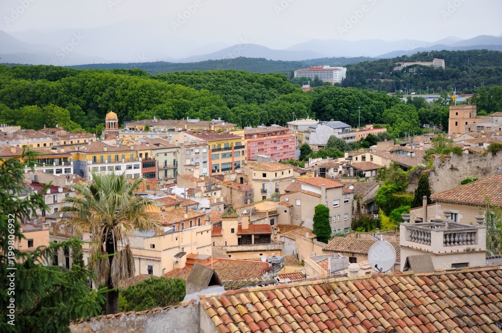 View of the city of Girona from the medieval pedestrian border wall. Roofs of houses, trees, mountains in a haze in the background. Cloudy sky. Girona, Spain