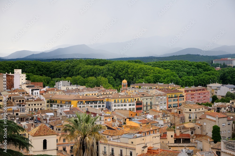 View of the city of Girona from the medieval pedestrian border wall. Roofs of houses, trees, mountains in a haze in the background. Cloudy sky. Girona, Spain