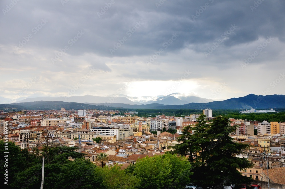 View of the city of Girona from the medieval pedestrian border wall. Roofs of houses, trees. Storm clouds over the city, somewhere sunlight makes its way through the clouds. Girona, Spain