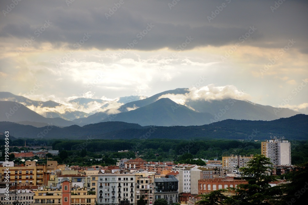 View of the city of Girona from the medieval pedestrian border wall. Roofs of houses, trees. Storm clouds over the city, somewhere sunlight makes its way through the clouds. Girona, Spain View of the city of Girona from the medieval pedestrian border wall. Roofs of houses, trees. Storm clouds over the city, somewhere sunlight makes its way through the clouds. Girona, Spain