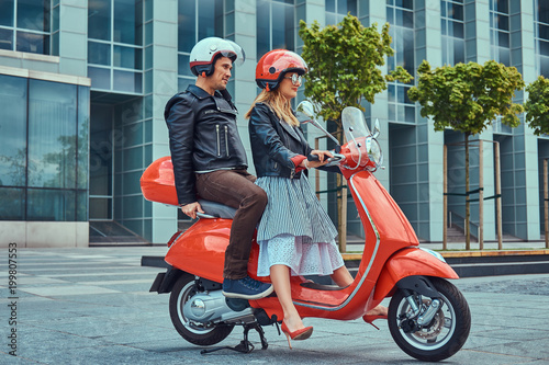 An attractive couple, a handsome man and sexy female riding together on a red retro scooter in a city. © Fxquadro