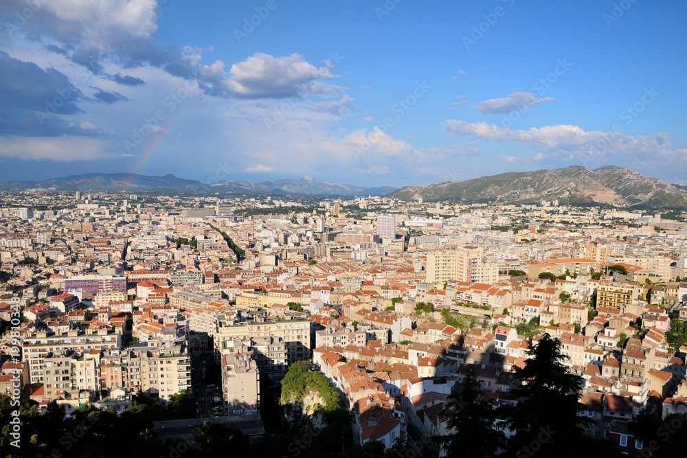 View from the height of the city.  Roofs of houses, trees, mountains in the background. Cloudy sky. A rainbow in the sky.