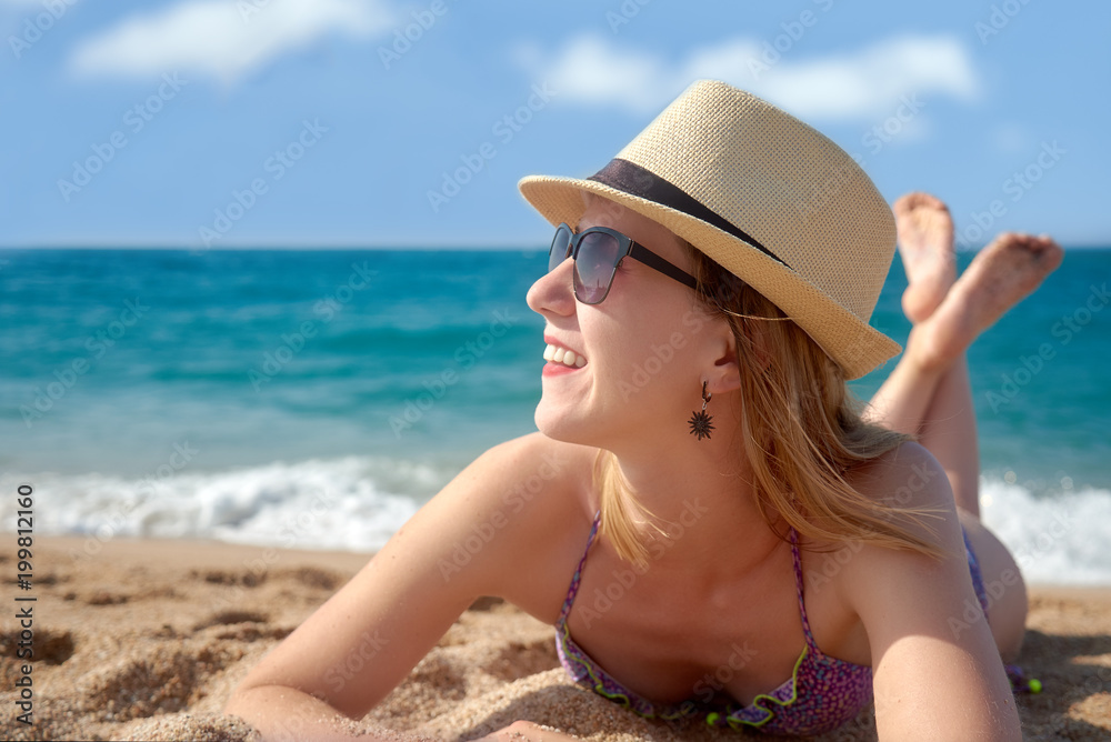 Young woman in sunhat and swimsuit is laying on the beach sand against the sea.