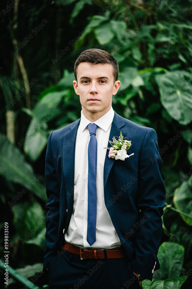 Serious handsome groom in a wedding suit and tie is standing on the background of greenery, on the jacket is a boutonniere of flowers. Man stands in the Botanical Garden full of greenery. Serious handsome groom in a wedding suit and tie is standing on the background of greenery, on the jacket is a boutonniere of flowers. Man stands in the Botanical Garden full of greenery.