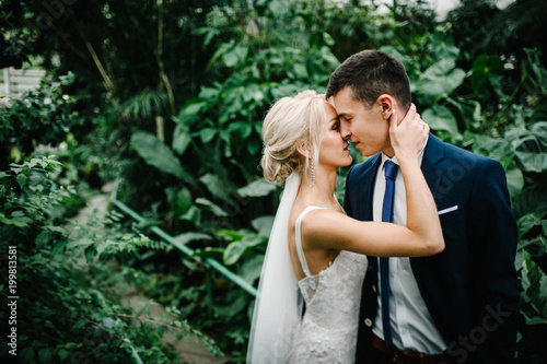 Portrait the groom in wedding suit and the bride in dress are standing and hugging, kissing in the Botanical green garden full of greenery. Wedding ceremony.