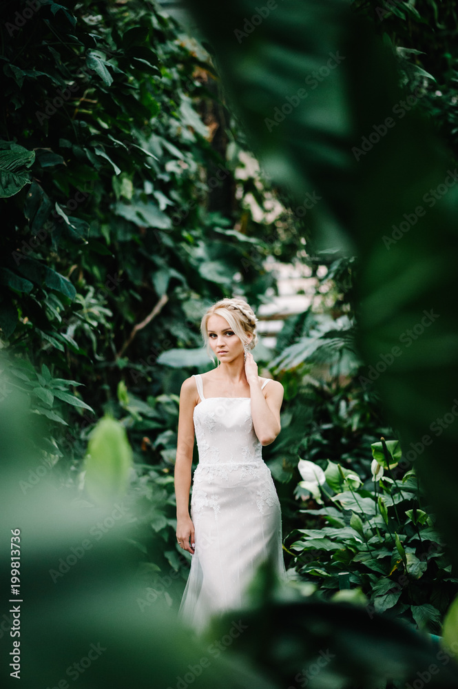 Woman stand in the Botanical green garden full of greenery. Wedding ceremony. Portrait attractive blonde bride standing in a wedding dress on the background of greenery. Woman stand in the Botanical green garden full of greenery. Wedding ceremony. Portrait attractive blonde bride standing in a wedding dress on the background of greenery.