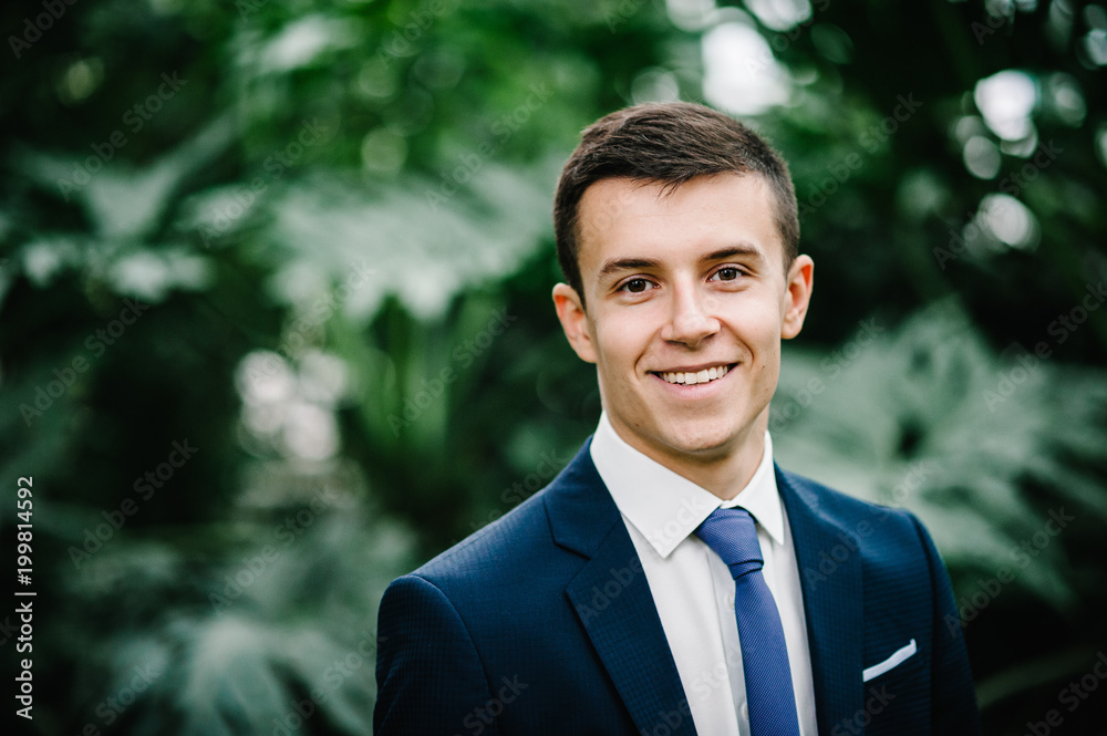 Happy portrait handsome groom in a wedding suit and tie is standing on the background of greenery. Man stands in the Botanical garden full of greenery. Happy portrait handsome groom in a wedding suit and tie is standing on the background of greenery. Man stands in the Botanical garden full of greenery.
