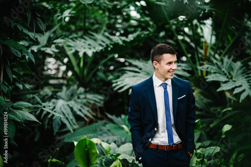 Happy portrait handsome groom in a wedding suit and tie is standing on the background of greenery. Man stands in the Botanical garden full of greenery. photo