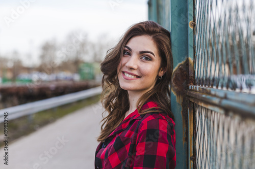 young beautiful woman standing by a fence. she is wearing casual clothes and smiling. Outdoors city background. Lifestyle